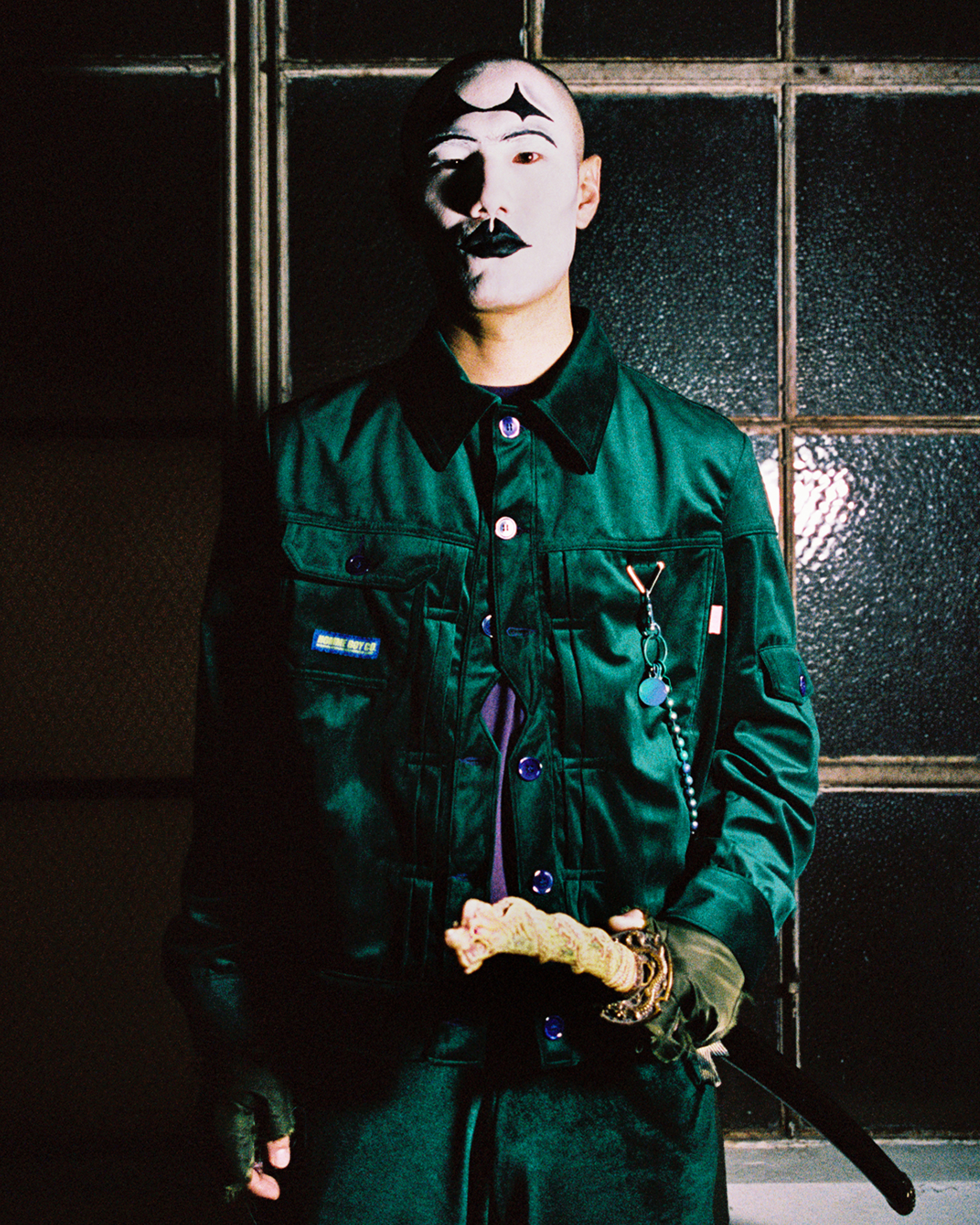 Model with black and white joker facepaint wearing a green ensemble with a shealthed sword on his hip